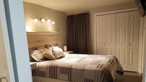 Quarto com cama grande, cabeceira, mesa de cabeceira, luminária, guarda-roupa e janela com cortinas.
