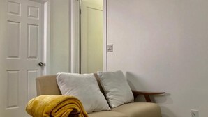 A neutral-colored sofa with two pillows and a yellow blanket in a room with white walls and a door. - Minimalist Living 1BR In-Law AC Unit with Private Washer & Dryer (Hayward, California)