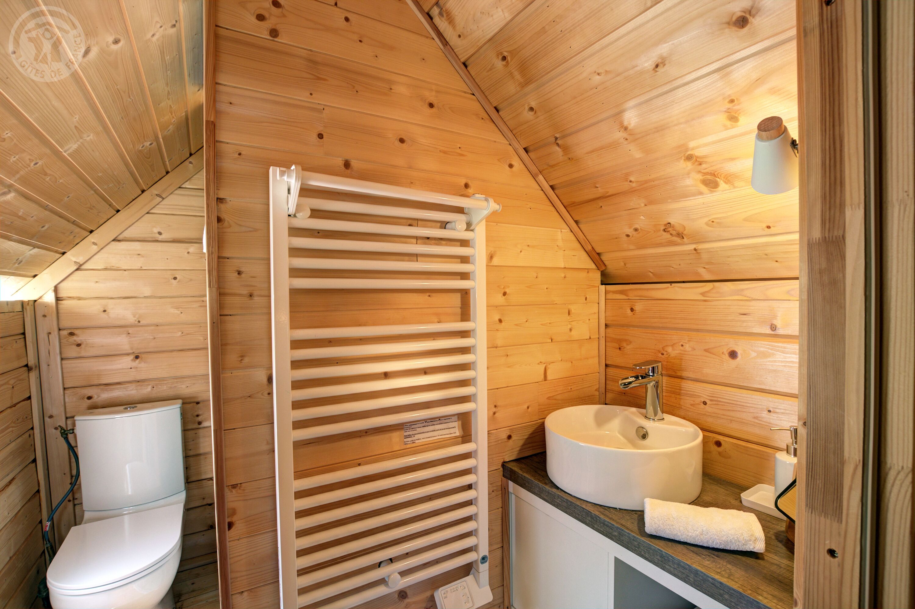 A compact wooden bathroom with a toilet, a shower with a white towel, and a sink with a mirror.
