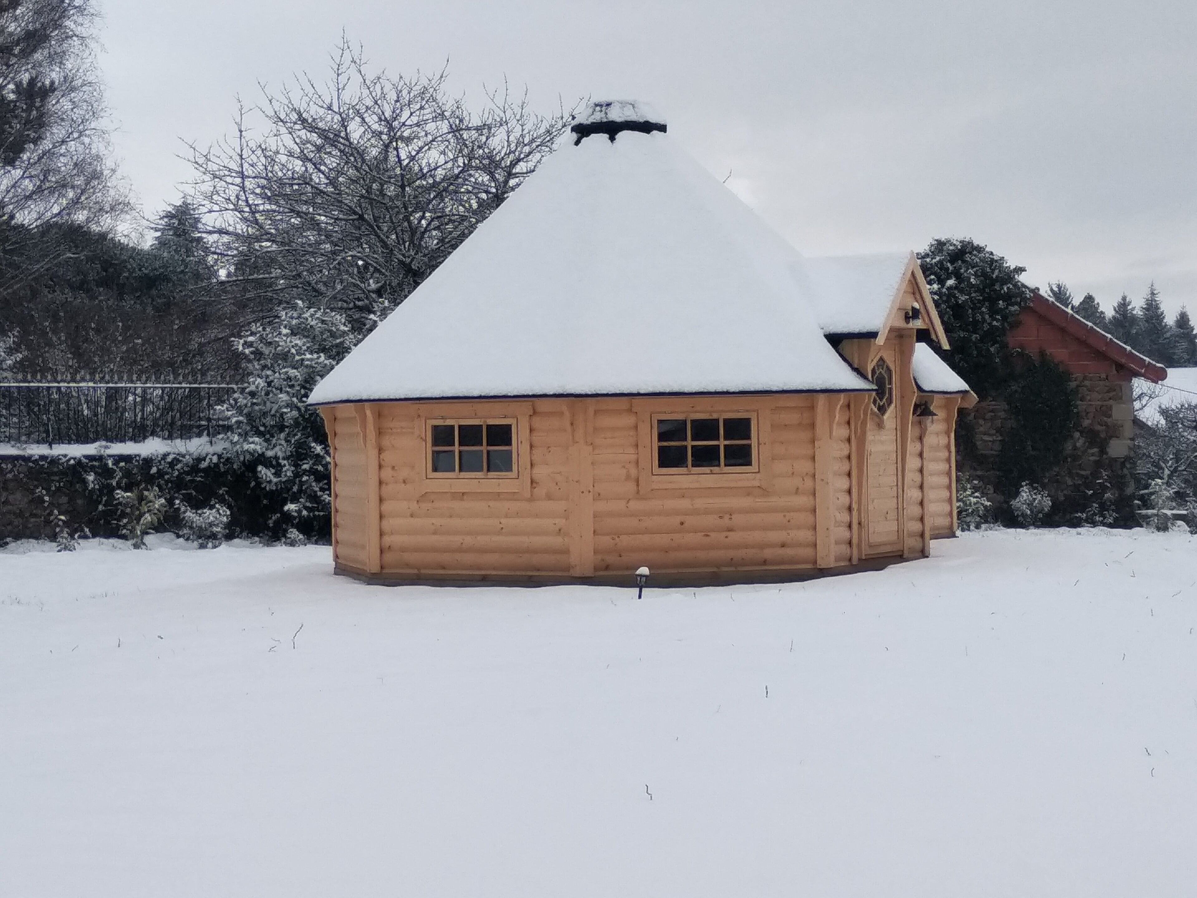 Una cabaña de madera con techo nevado, dos ventanas y una chimenea.