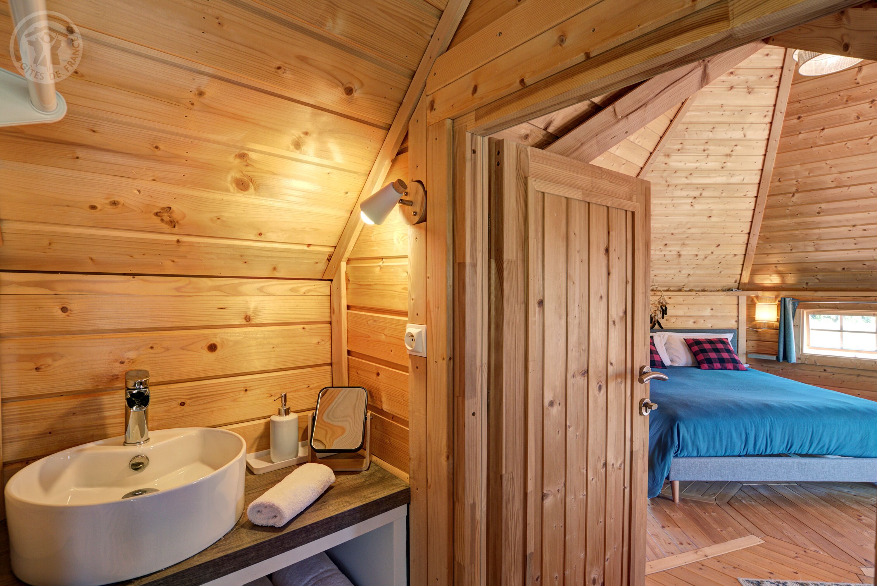 A wooden cabin interior with a bathroom featuring a sink, mirror, and towel. A bed with blue bedding is visible in the adjacent room.