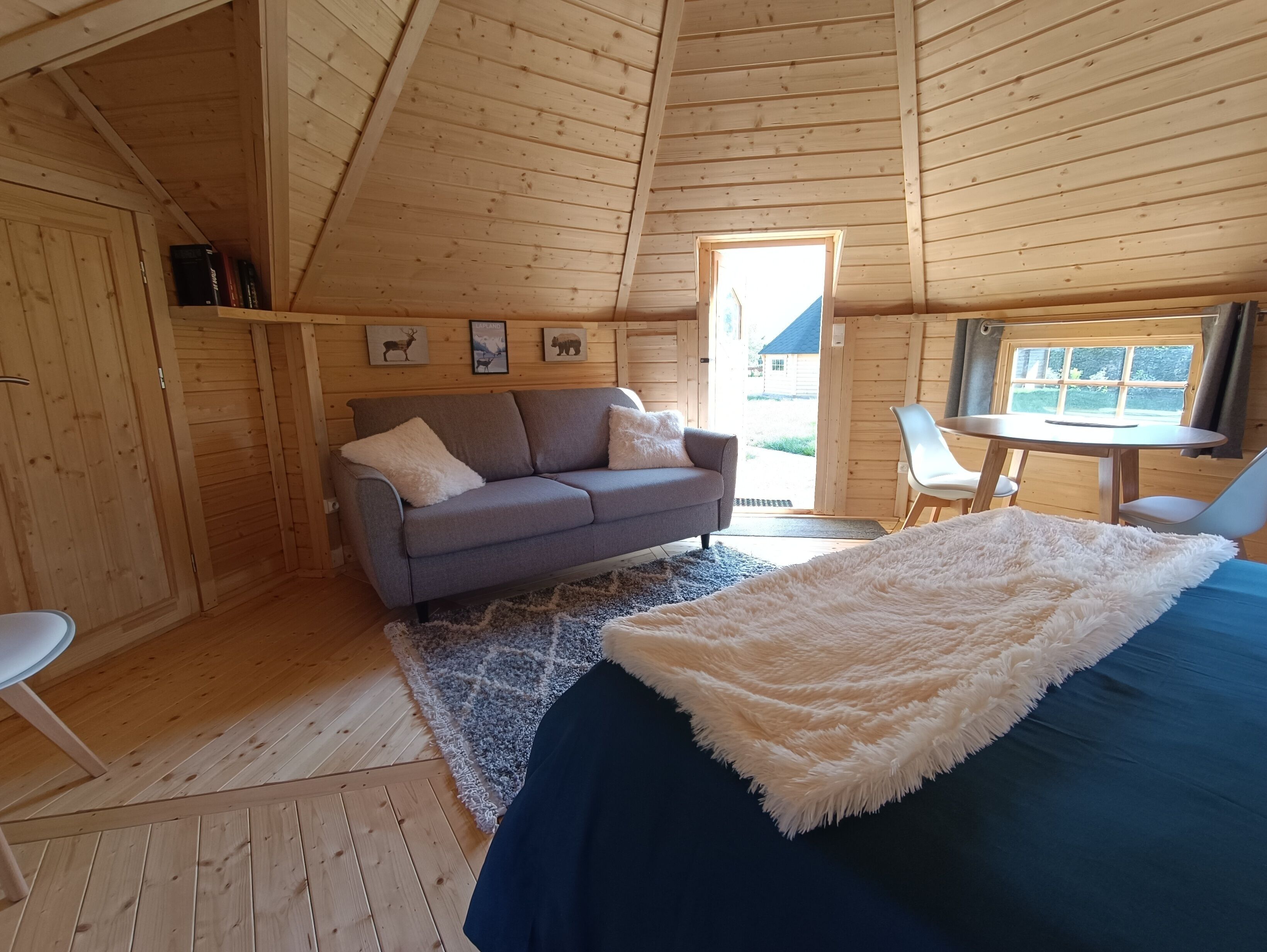 A cozy cabin interior with a sofa, a bed with a fluffy white blanket, a small table, and a window with a view of the outside.
