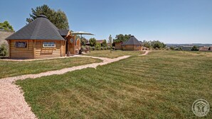 Une cabane en bois, accessible par un chemin de gravier, entourée d’un champ herbeux et d’arbres.