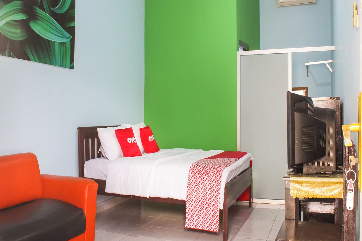 Quarto com uma cama, televisão, travesseiro vermelho e branco, parede verde e uma cadeira vermelha.