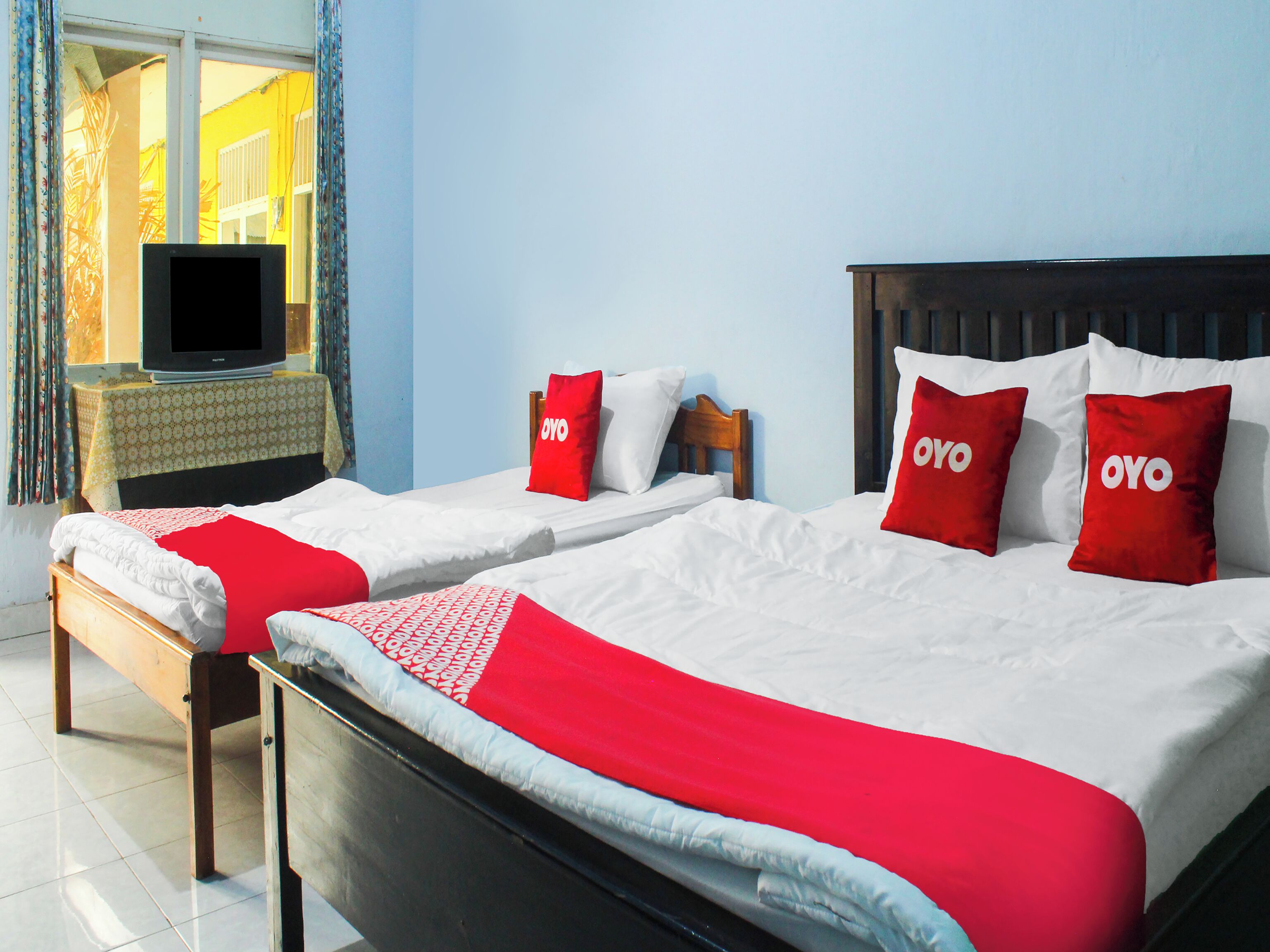 Una habitación de hotel con dos camas, cada una con una manta roja y blanca, un cabecero de madera y un televisor sobre una mesita.