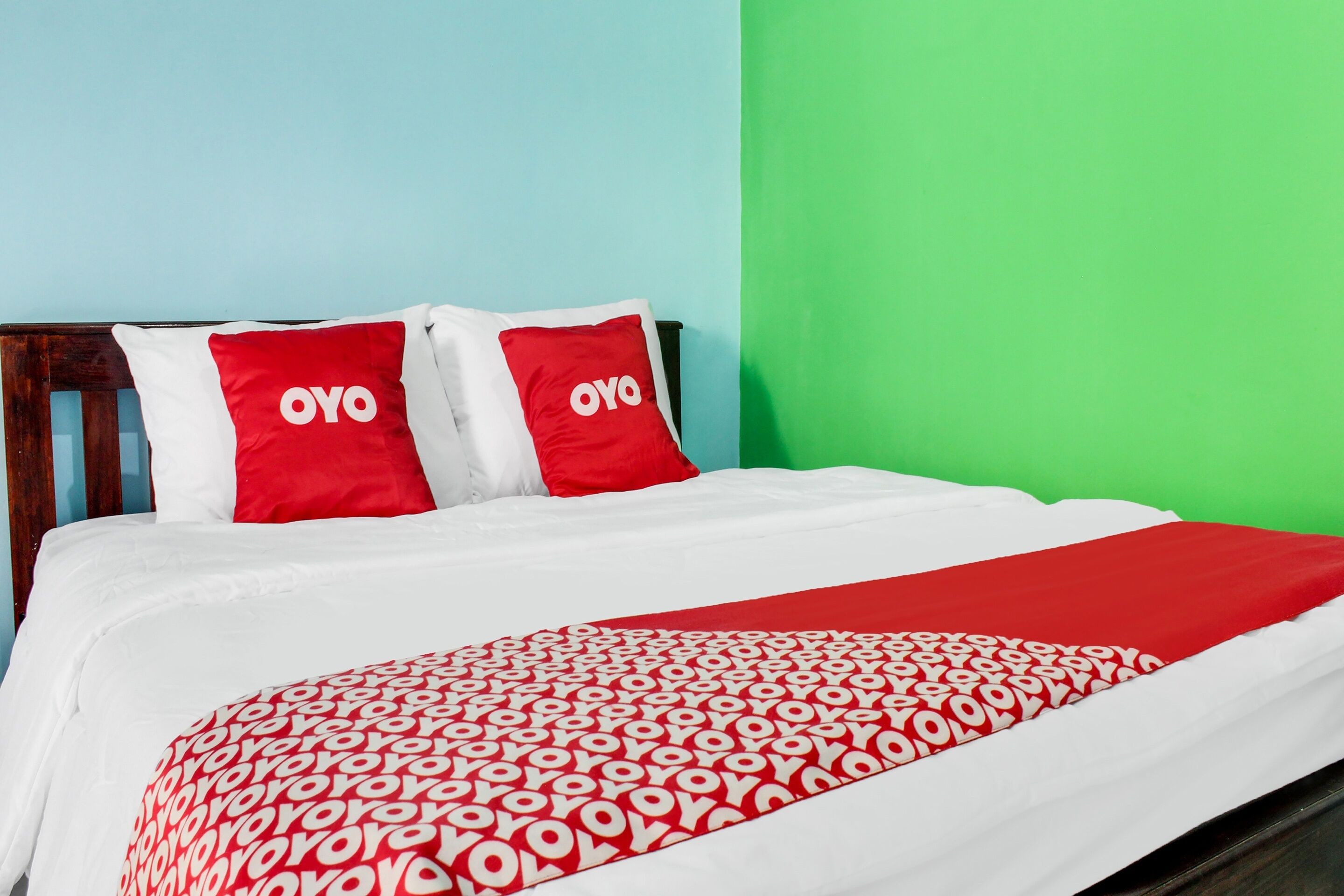 Een bed met een wit en rode spreideken, twee rode kussens met de tekst 'OYO' en een groene muur.