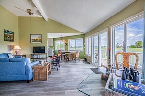 A spacious living room with a large window overlooking a golf course, a dining area with wicker furniture, and a cozy seating area with a blue sofa and a lamp. - Pets Considered, Golf Cart included - The Bird House (Fripp Island)