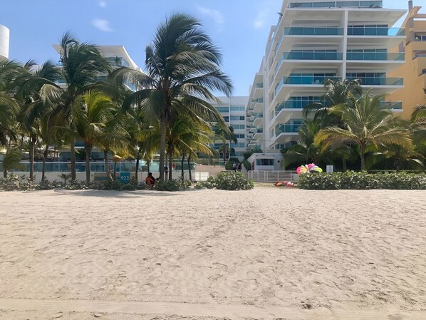 A sandy beach with palm trees and a modern building in the background.