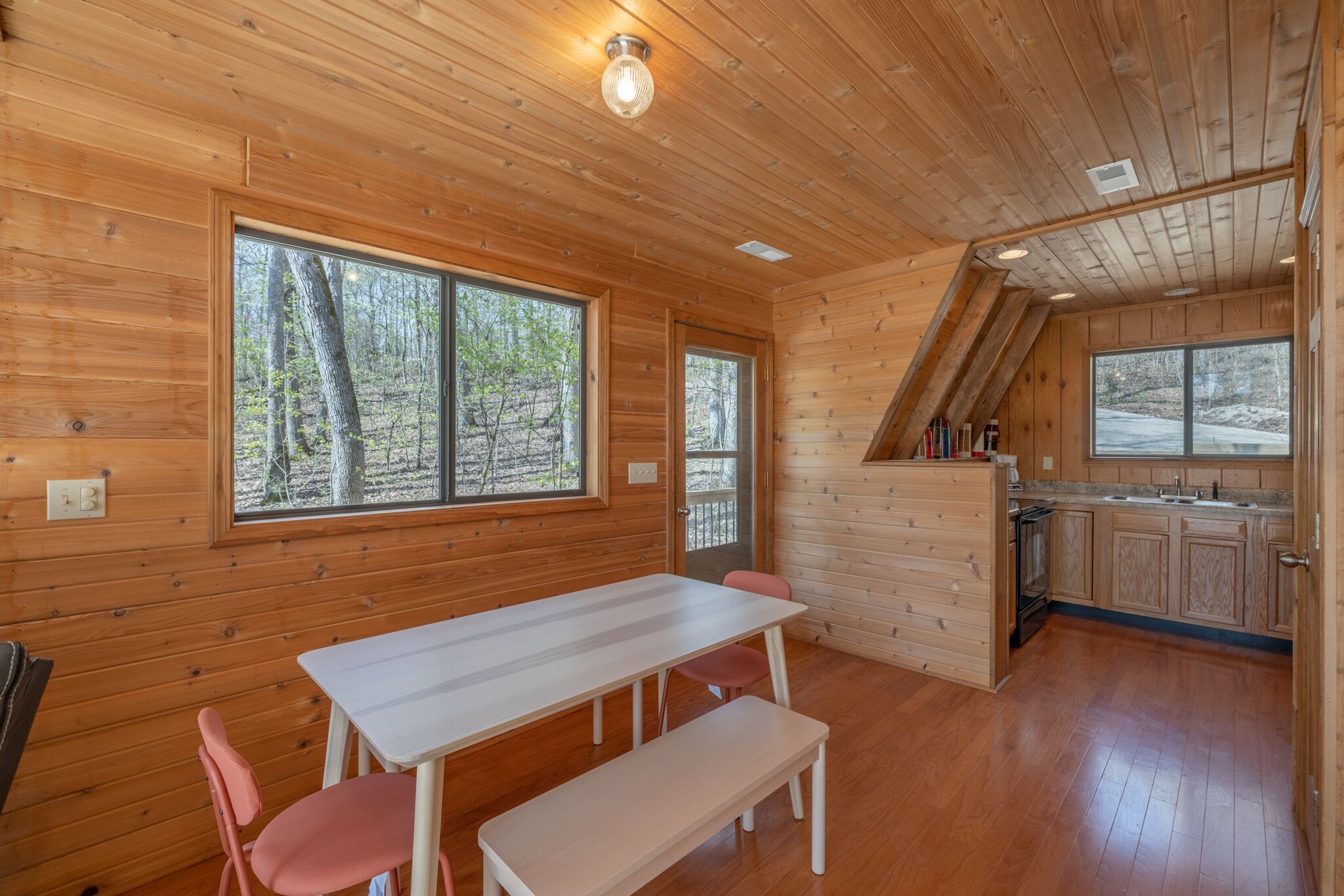 Una cabaña de madera con comedor, cocina y grandes ventanales que dan a un bosque.