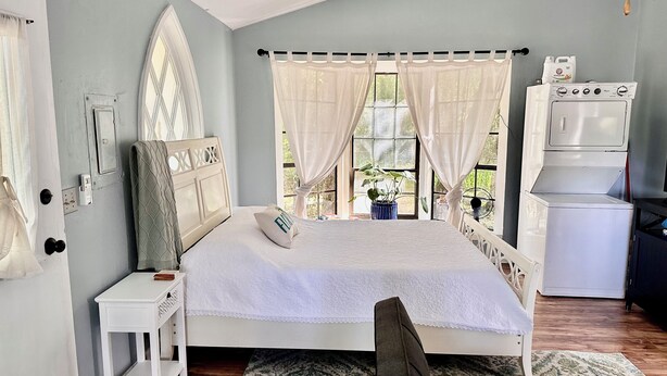 Un dormitorio con cama, mesita de noche, cortinas, una ventana con vistas, lavadora y ventilador.