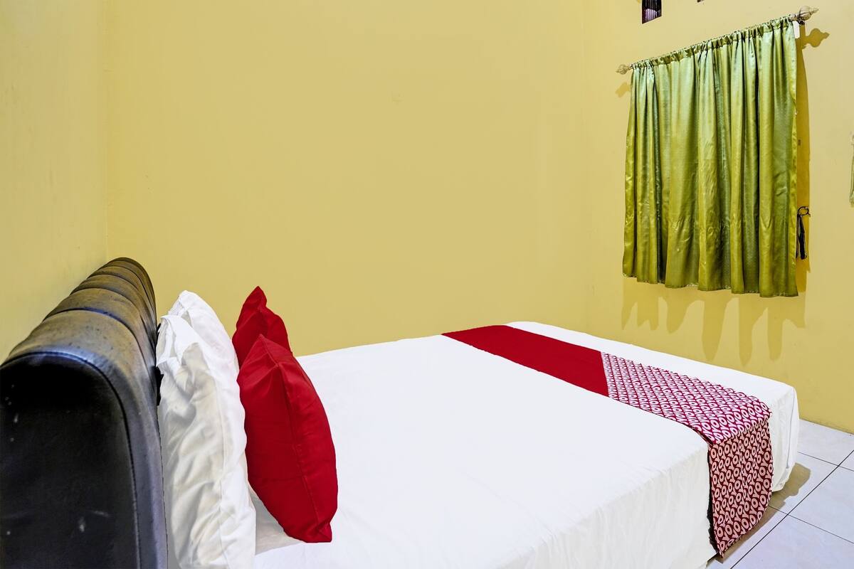 Ein Zimmer mit einem Bett, einem roten Kissen, einem weißen Kissen, einem grünen Vorhang und einem schwarzen Gegenstand auf dem Boden.