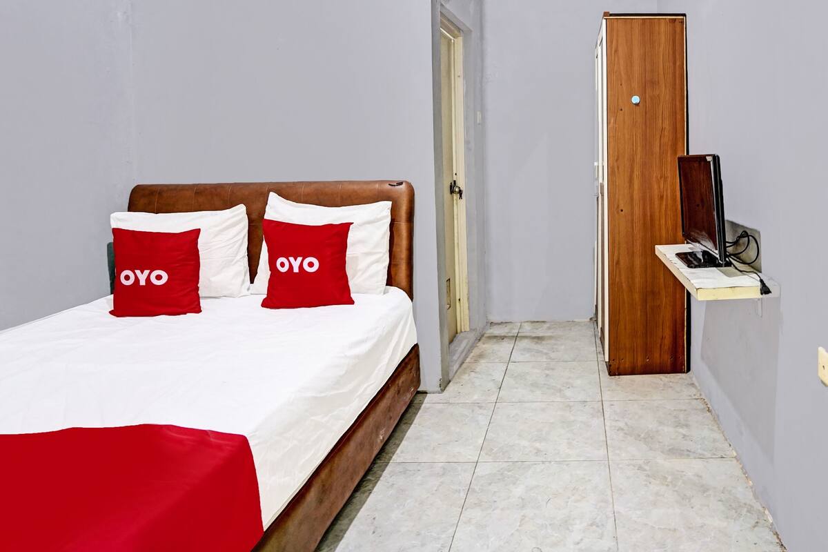Ein ordentlich bezogenes Bett mit weißer und roter Bettdecke, zwei rote Kissen mit dem Aufdruck "OYO", ein Holz-Kopfbrett und eine kleine Holzablage mit einem Laptop.