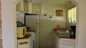 Eine schmale Küche mit weißen Schränken, einer Mikrowelle und einem Kühlschrank. Ein Fenster mit Vorhängen und einer Wandleuchte sind vorhanden.