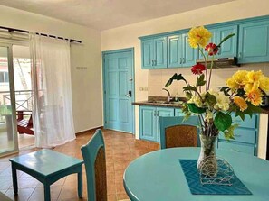Een kamer met een blauwe tafel, een stoel, een vaas met bloemen en een keukenhoek met blauwe kastjes.