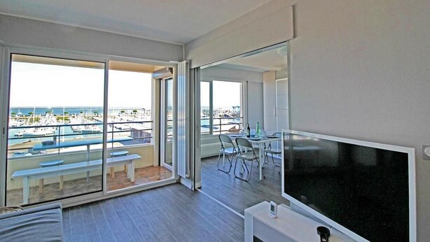 A modern room with a large TV, a balcony with a view of a marina, and a dining area with chairs and a table. - L'abricôtier - Arcachon (ARCACHON)