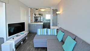 A modern living room with a flat-screen TV, grey sofa, striped pillows, and a kitchen in the background. - L'abricôtier - Arcachon (ARCACHON)