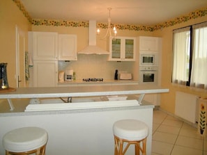 A kitchen with white cabinets, a central island with stools, a chandelier, and a window with curtains. - Gite LE Logis DE LA Perichere (CASTRES-GIRONDE)