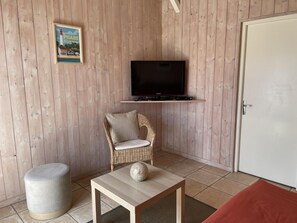Een kamer met een aan een houten muur bevestigde televisie, een rieten stoel met kussens, een klein rond tafeltje met een decoratieve bal, en een witte deur.
