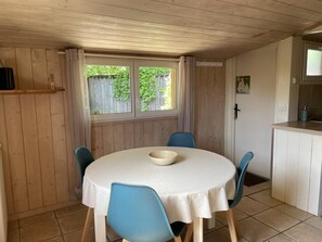 A room with a round table, blue chairs, a white tablecloth, a window with curtains, and a kitchen area.