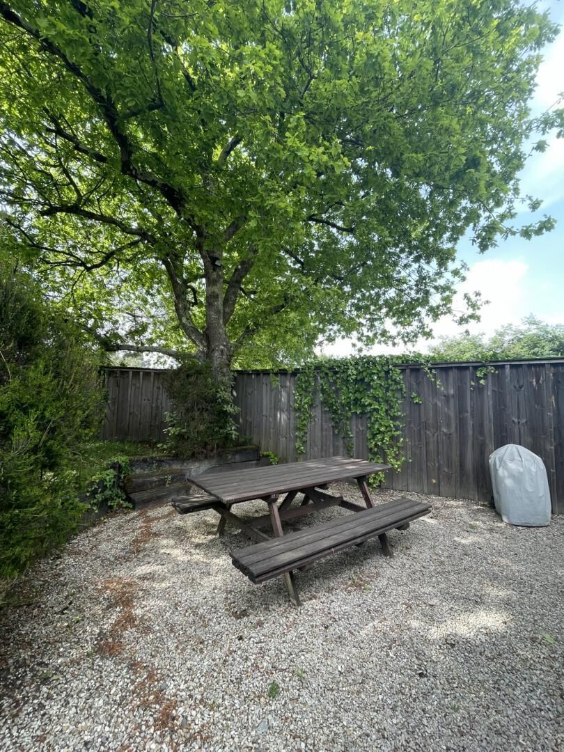 A wooden picnic table with benches in a gravel area, surrounded by a wooden fence and greenery.