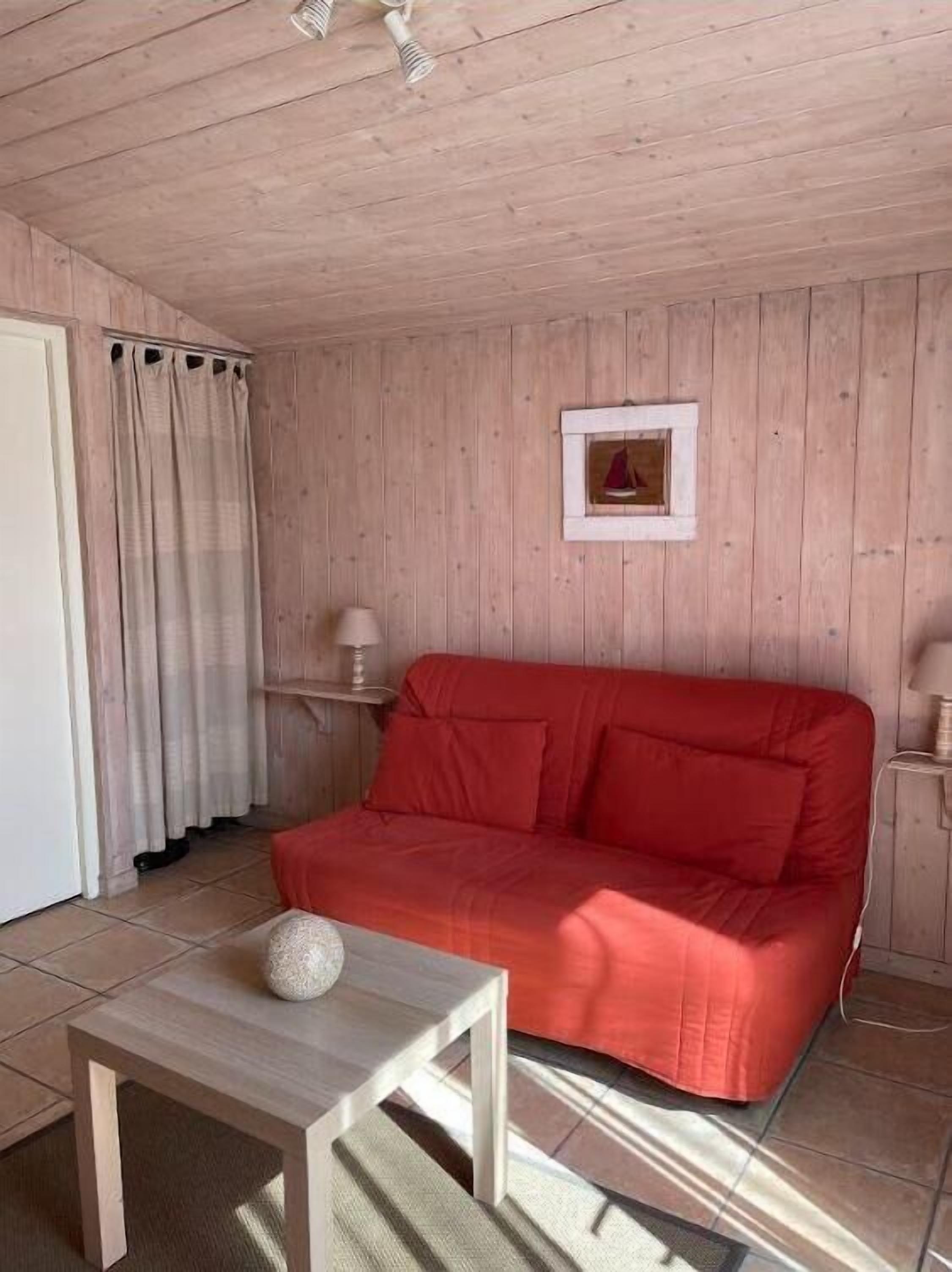 Una habitación con paredes de madera, un sofá rojo, una mesita con un objeto decorativo, un cuadro enmarcado en la pared y una ventana con cortinas.