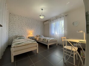 A room with two single beds, a desk with a chair, a small table, and a ceiling light.