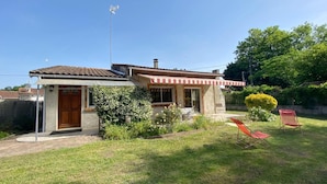 Ein Haus mit kleiner Garage, einer roten und weißen Markise sowie einem Garten mit Rasen und zwei roten Stühlen.