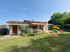Ein Haus mit kleiner Garage, einer roten und weißen Markise sowie einem Garten mit Rasen und zwei roten Stühlen.