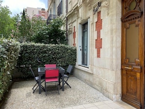 Een terras buiten met een tafel, stoelen, een haag en een gevel met een deur waarop 'Dacha' staat.