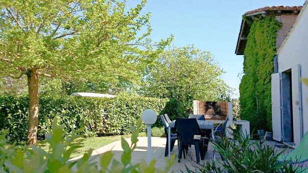 Un patio con una mesa y sillas, rodeado de follaje y un edificio con techo de tejas.