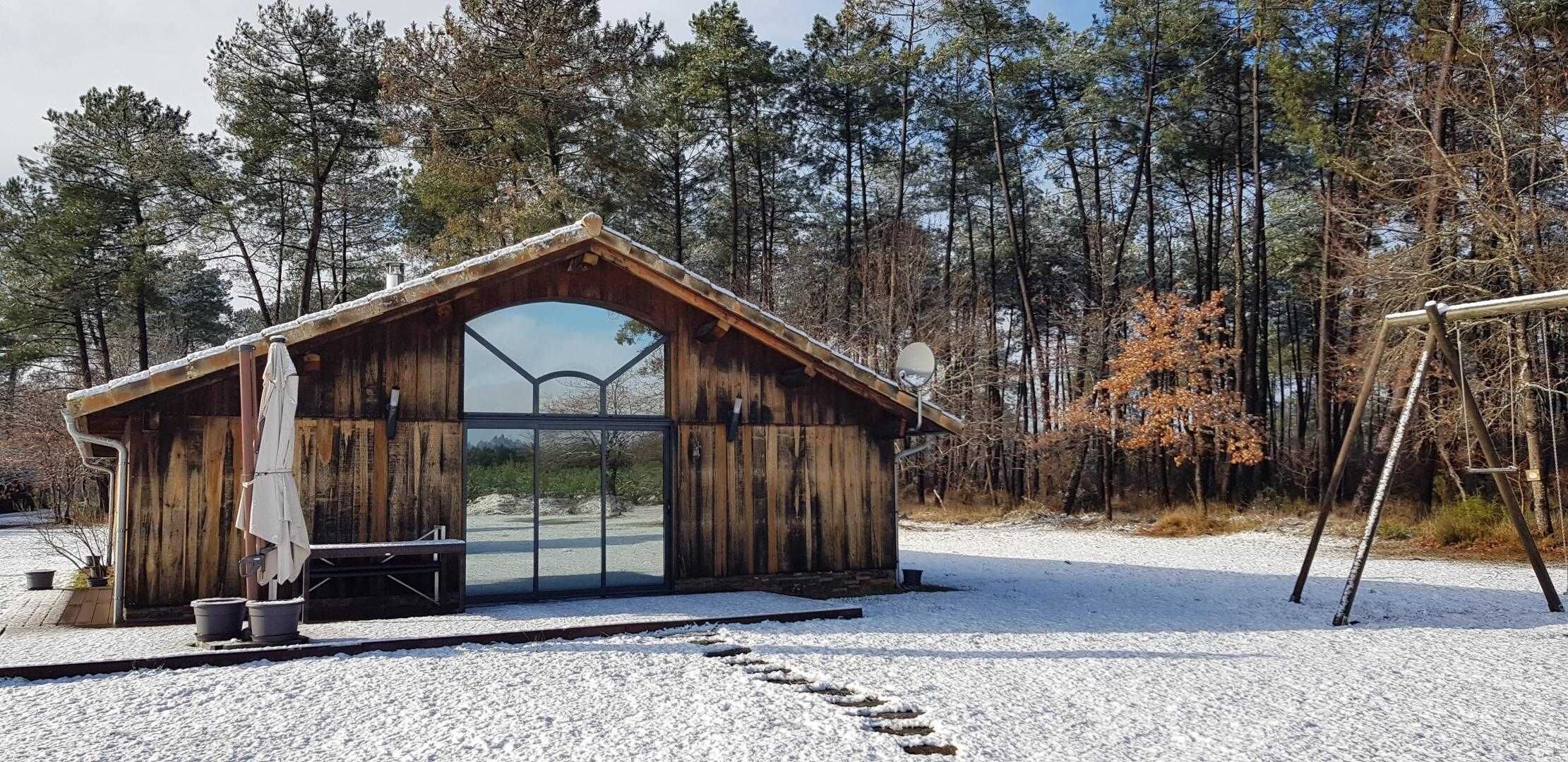 A wooden cabin with a large glass door, surrounded by a snowy landscape and trees.