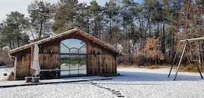 A wooden cabin with a large glass door, surrounded by a snowy landscape and trees.