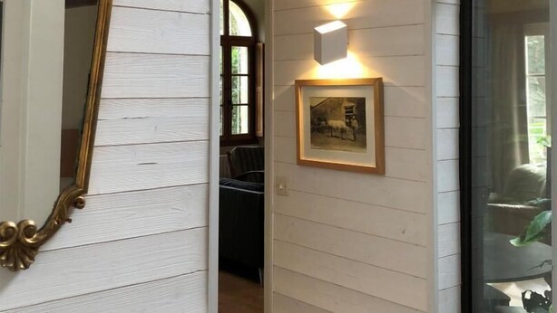 A hallway with white wooden walls, a framed mirror, a wall-mounted light, and a framed picture.