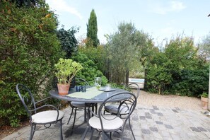 An outdoor patio with a round table and four chairs, surrounded by dense greenery and a gravel path.