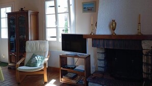 Ein Wohnzimmer mit Holzboden, einem Fernseher auf einem Schrank, einem Kamin, einem Bücherregal und einem Fenster mit Vorhängen.