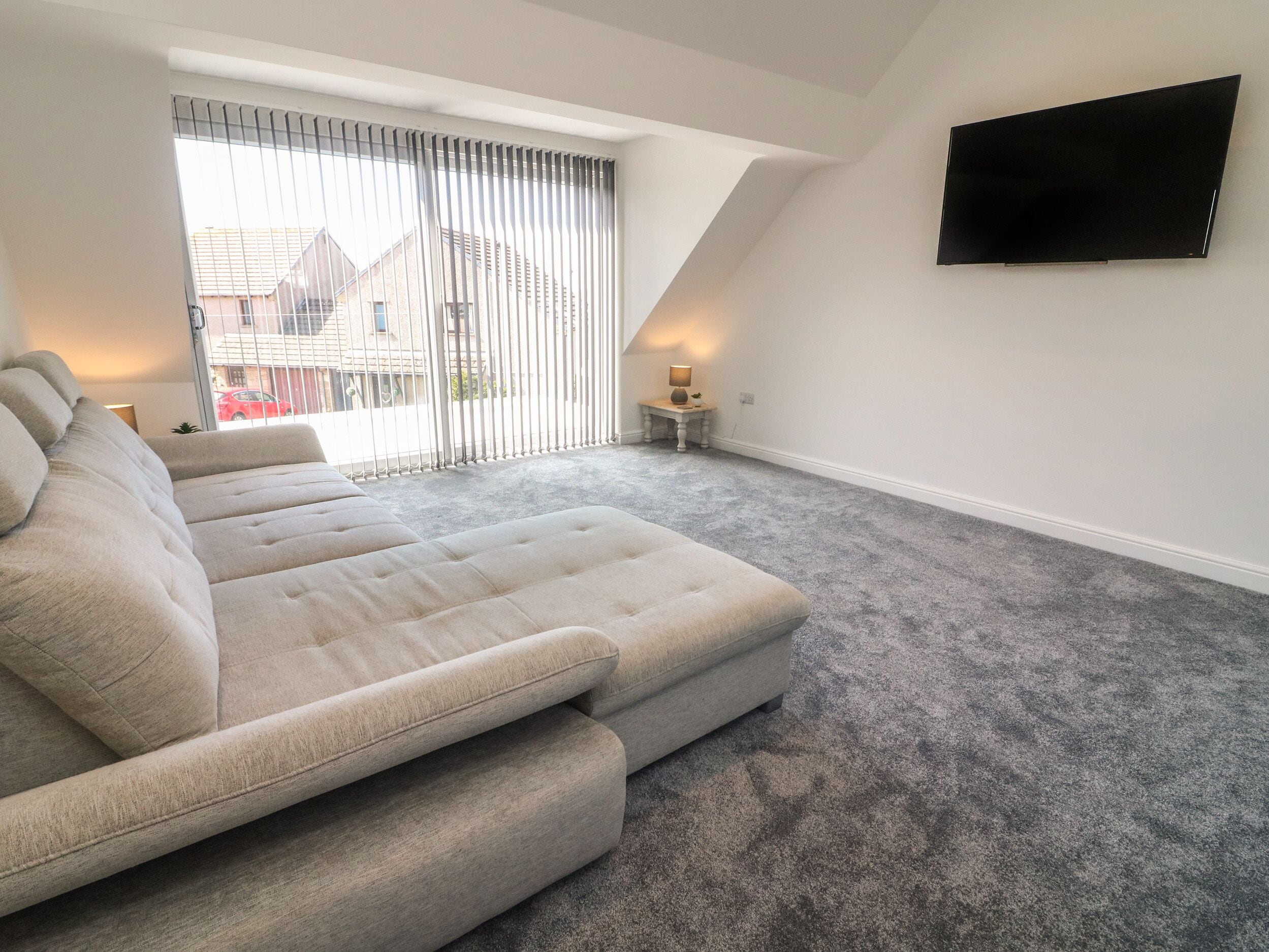 A modern living room with a sectional sofa, a flat-screen TV mounted on the wall, and a large window with blinds.