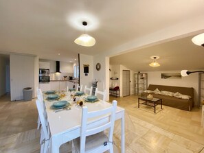 A dining area with a white table set for four, a kitchen in the background, and a living room with a sofa and a coffee table.