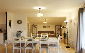Un coin repas avec une table et des chaises blanches, un lustre et une vue sur un salon comprenant un canapé et une plante.