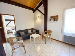 Een kamer met een donkergrijze bank, twee rieten fauteuils, een witte salontafel en een boekenkast vol boeken.