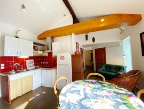 Une cuisine compacte avec des meubles blancs, un plan de travail carrelé de couleur rouge, un réfrigérateur blanc et un coin repas agrémenté d’une nappe à motifs floraux.