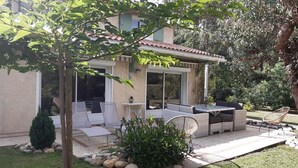 Una casa con patio arredato, una zona relax coperta e un giardino con un piccolo albero e piante.