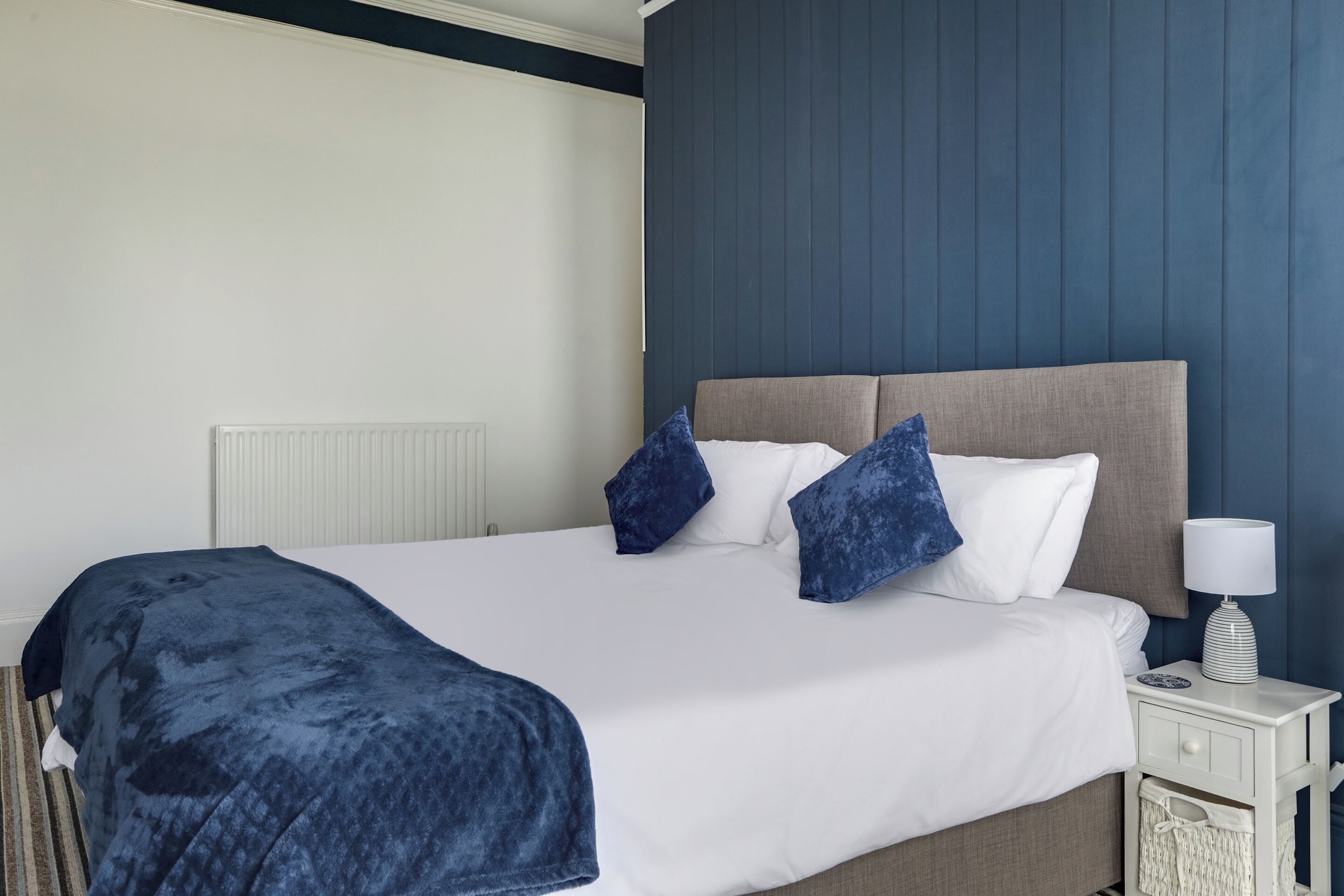 A neatly made bed with a white comforter and blue pillows, a bedside lamp, and a small bedside table with a lamp.