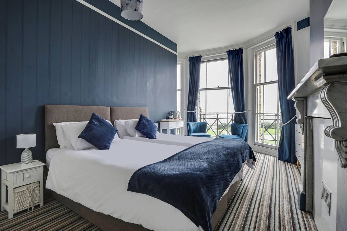 A hotel room with a large bed, a nightstand, a chair, and a view of the outdoors through large windows.