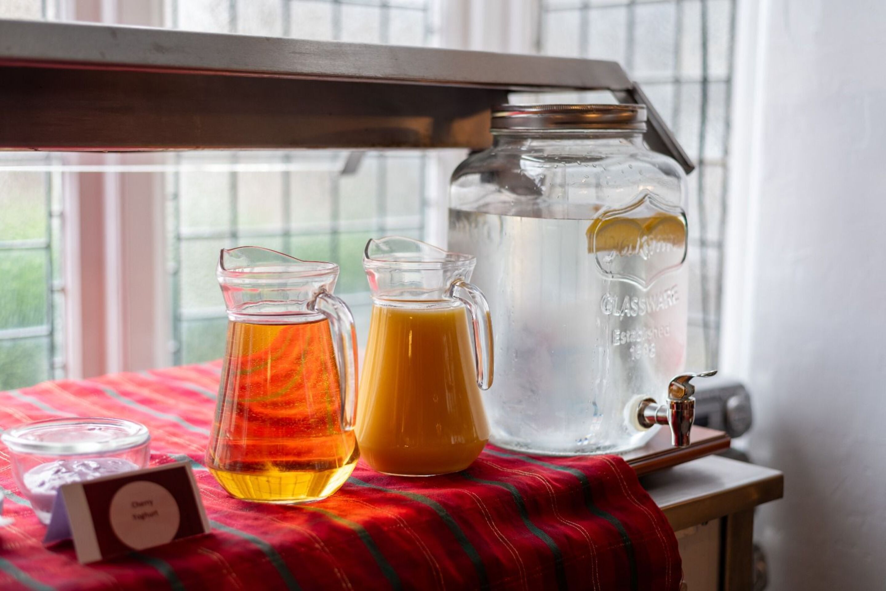 A table with a pitcher of iced tea, a glass of orange juice, and a large glass jar of water.