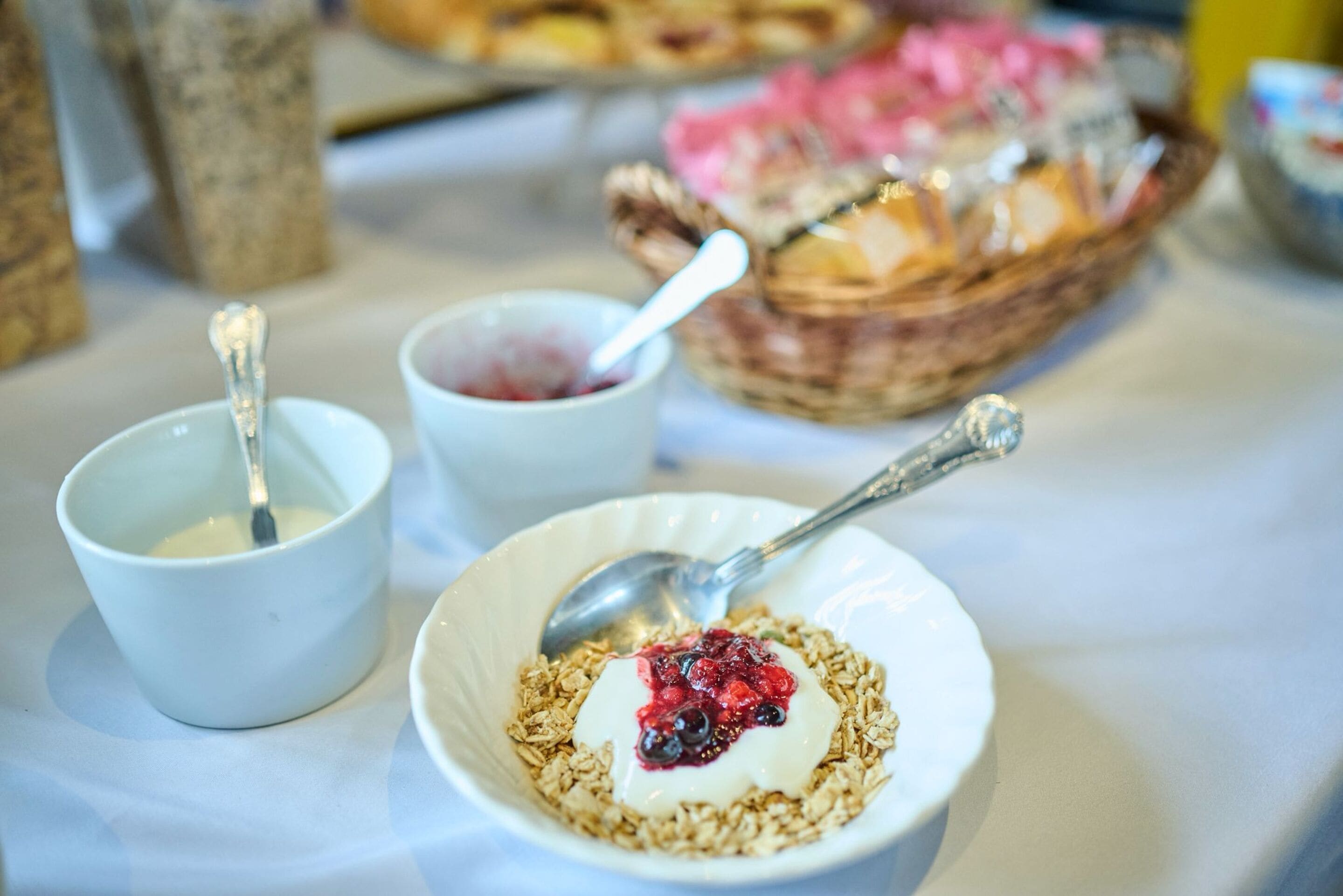 En frukostuppsättning med skålar fyllda med yoghurt och müsli, en sked och en korg med bakverk.