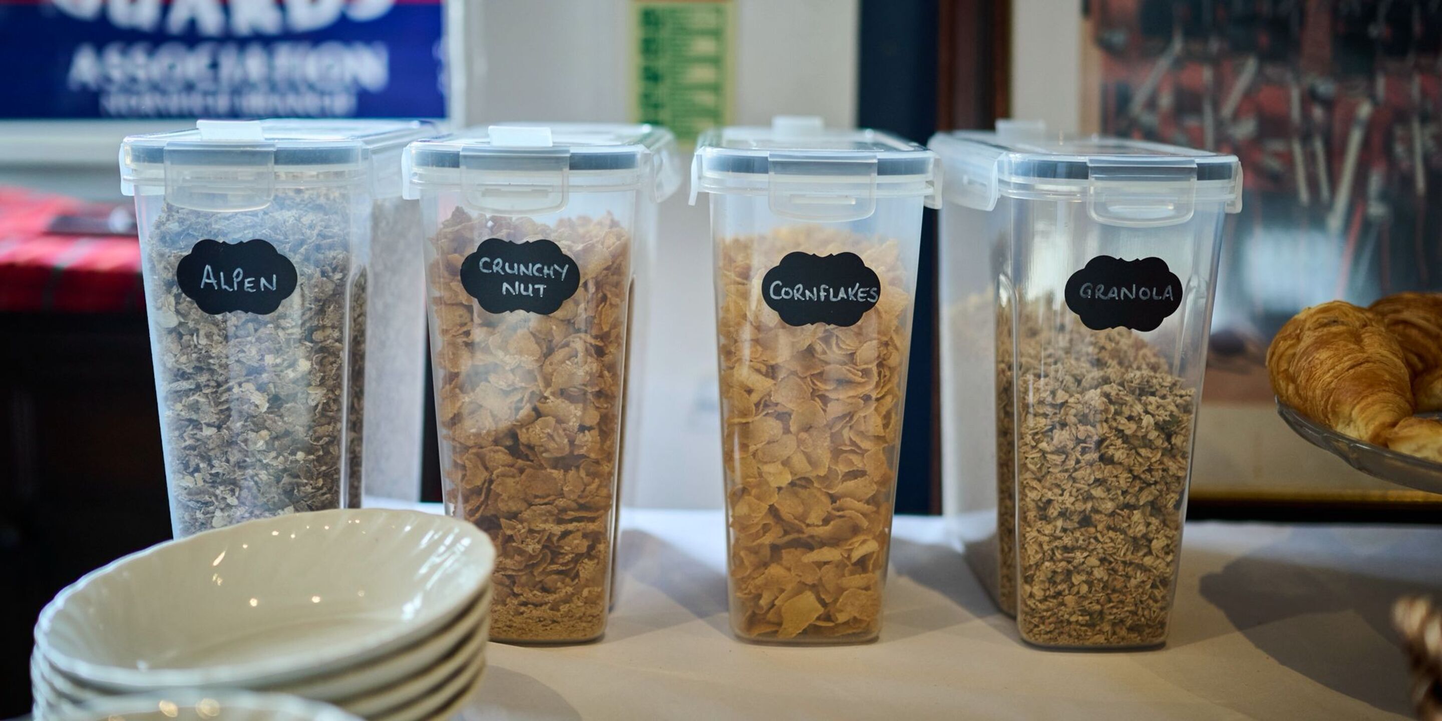 Vier gemarkeerde bakken met granola, cornflakes en andere ontbijtgranen op een tafel.