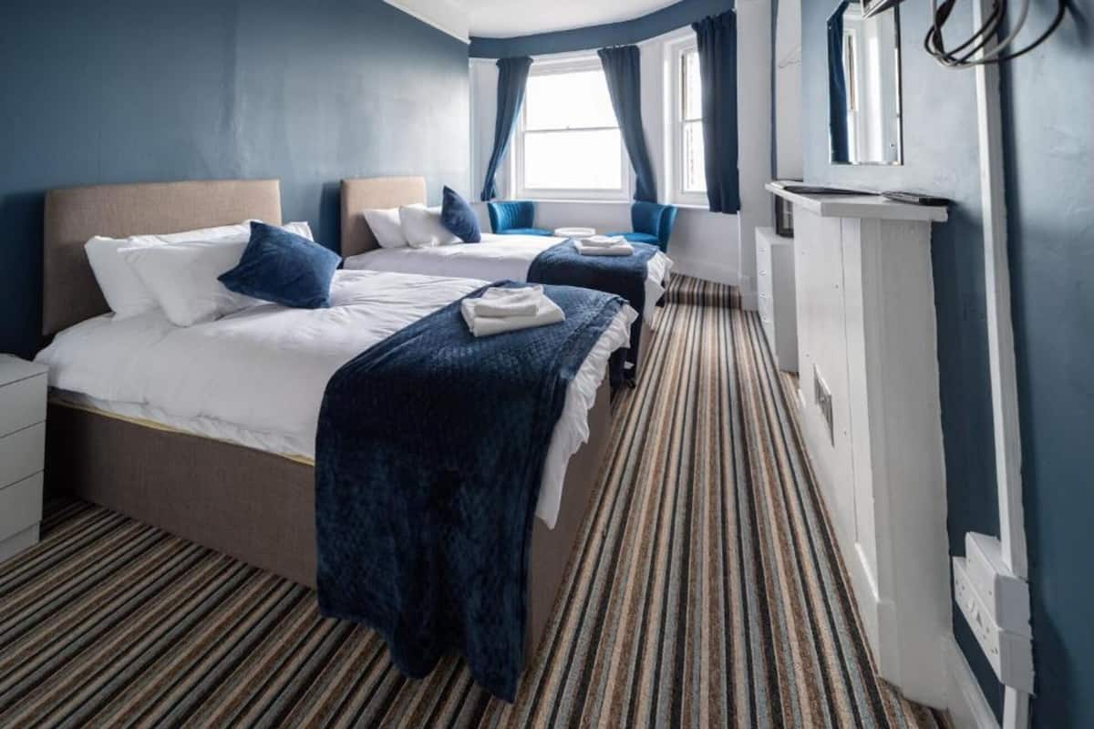 A hotel room with two beds, a large window, a bedside table, and a striped carpet.