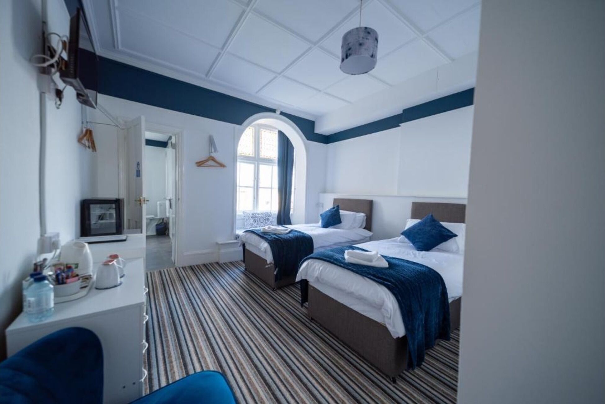 Habitación de hotel con dos camas, un ventanal arqueado grande, alfombra a rayas y una lámpara colgante.
