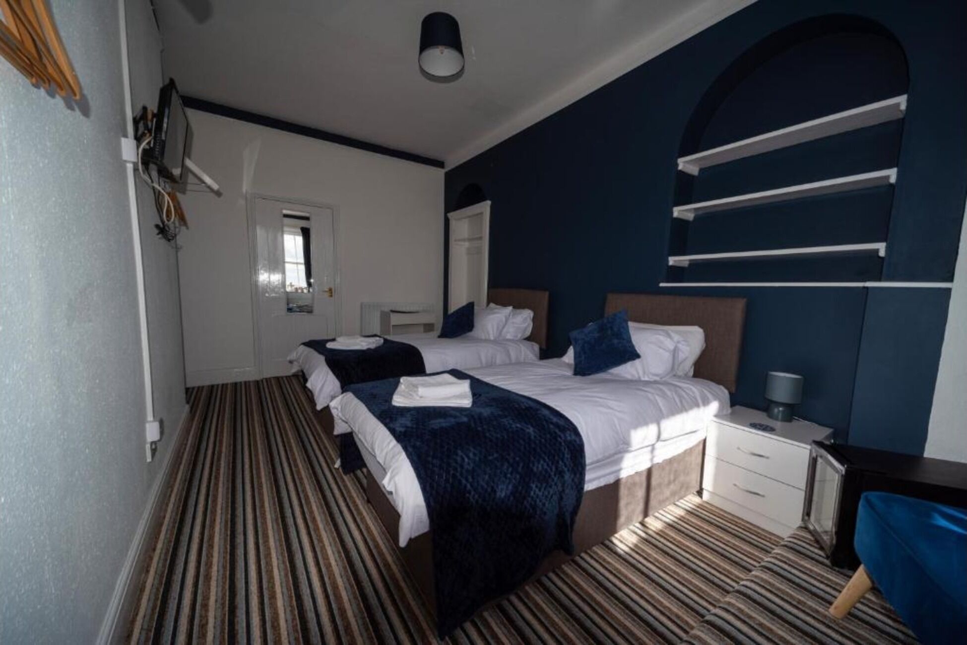 Ein Hotelzimmer mit zwei Betten, einem großen Spiegel, einem Fenster mit Jalousien, einem Nachttisch mit Lampe und einem Streifenmuster-Teppich.
