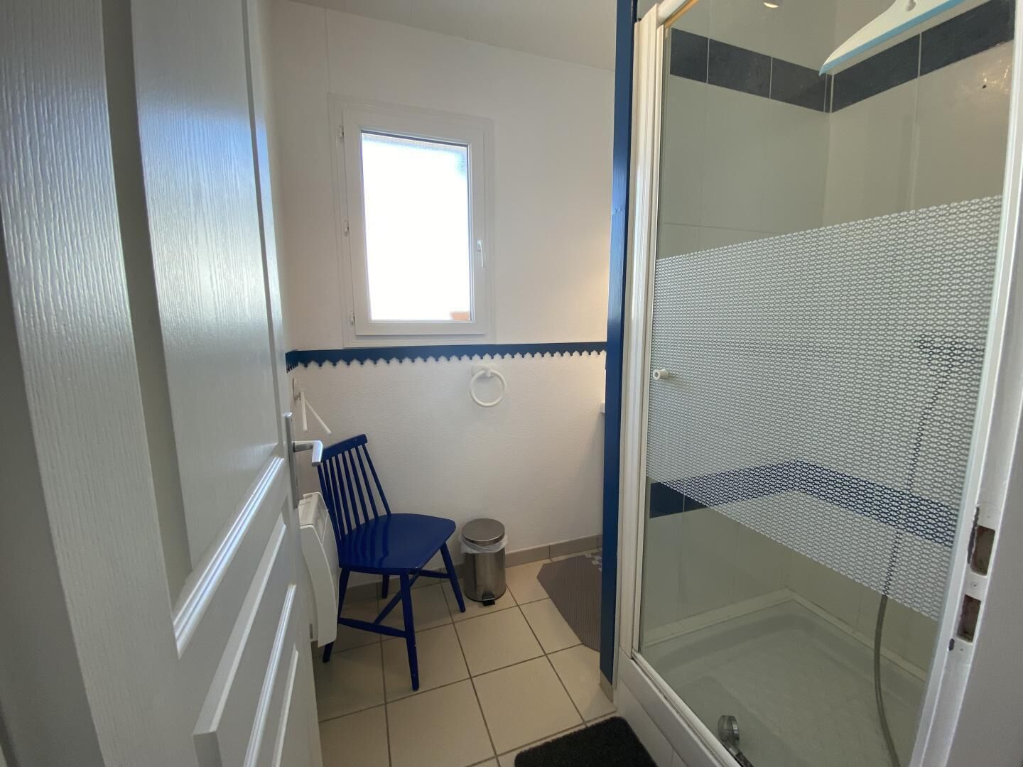 Une salle de bain avec une douche, une fenêtre, une chaise et une poubelle.